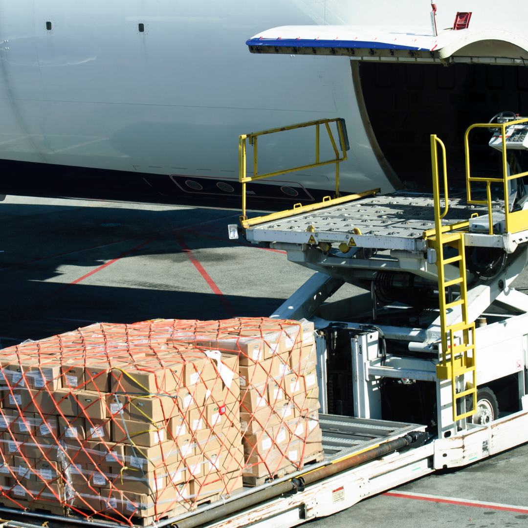Cargo boxes strapped on a conveyor cart next to a cargo plane at the gate, with a yellow lift platform and open aircraft cargo door visible.